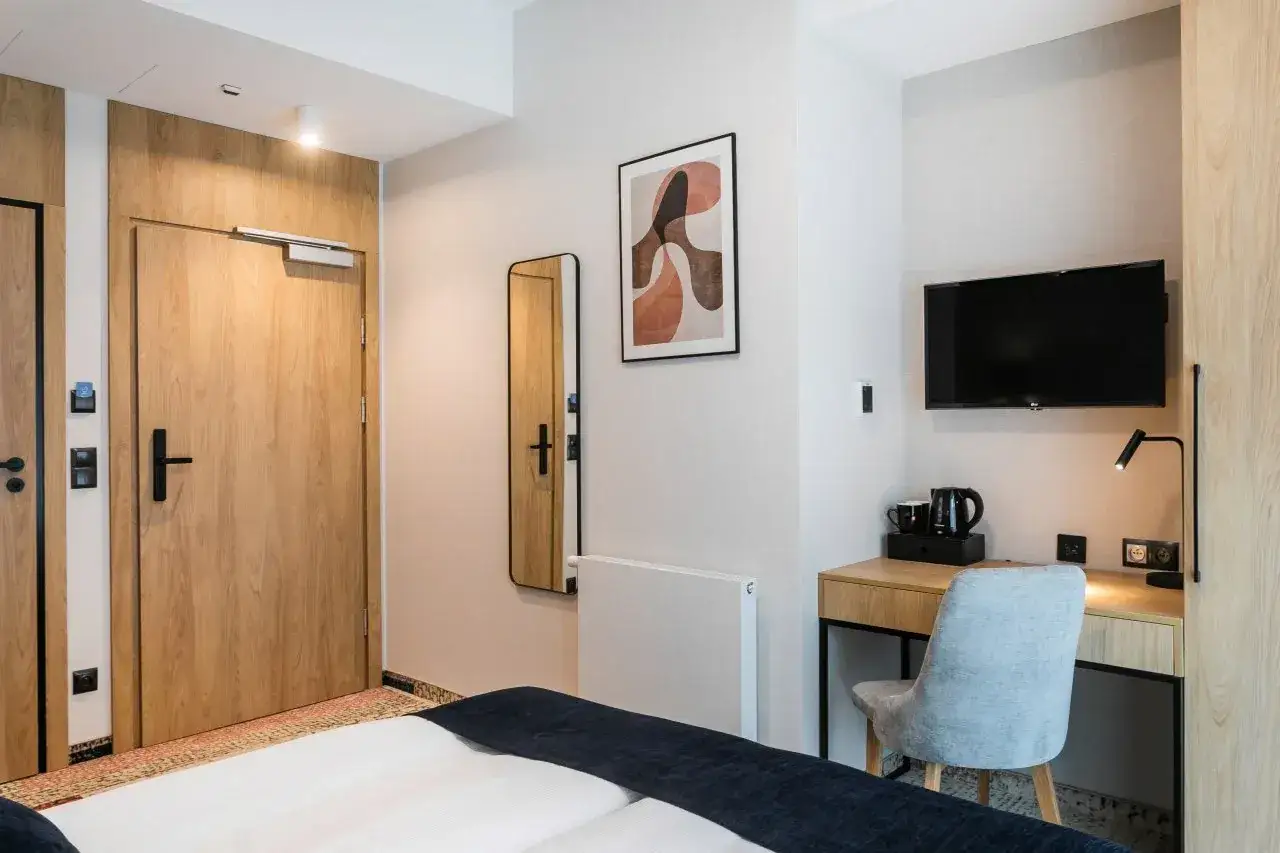 Pok&oacute;j w hotelu Vesper House: ł&oacute;żko, biurko z czajnikiem i TV, lustro, obraz i drewniane drzwi.