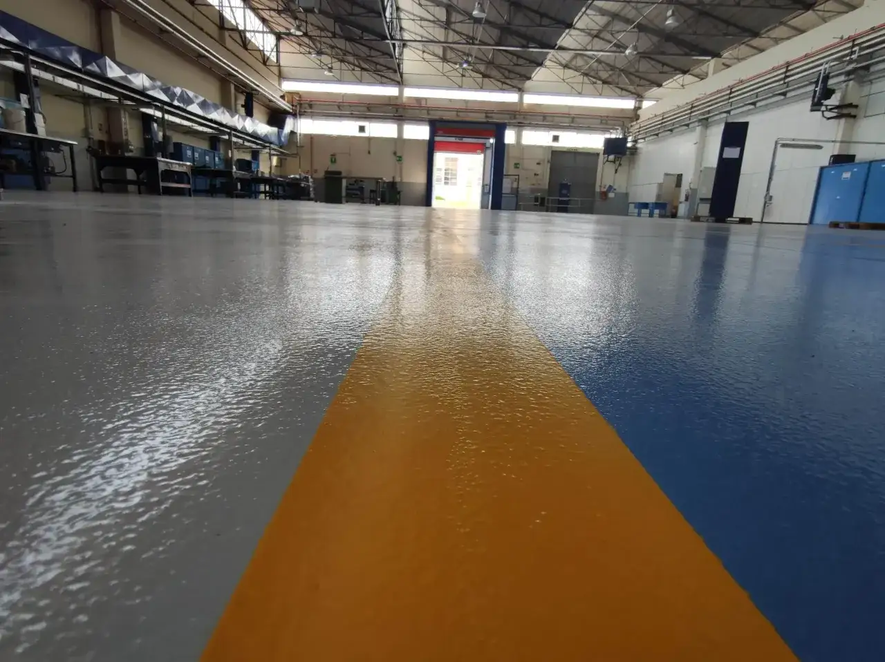 Pavimento industriale appena finito, con finitura lucida in resina e segnaletica a fasce gialla e blu. Pronto per lavorare.