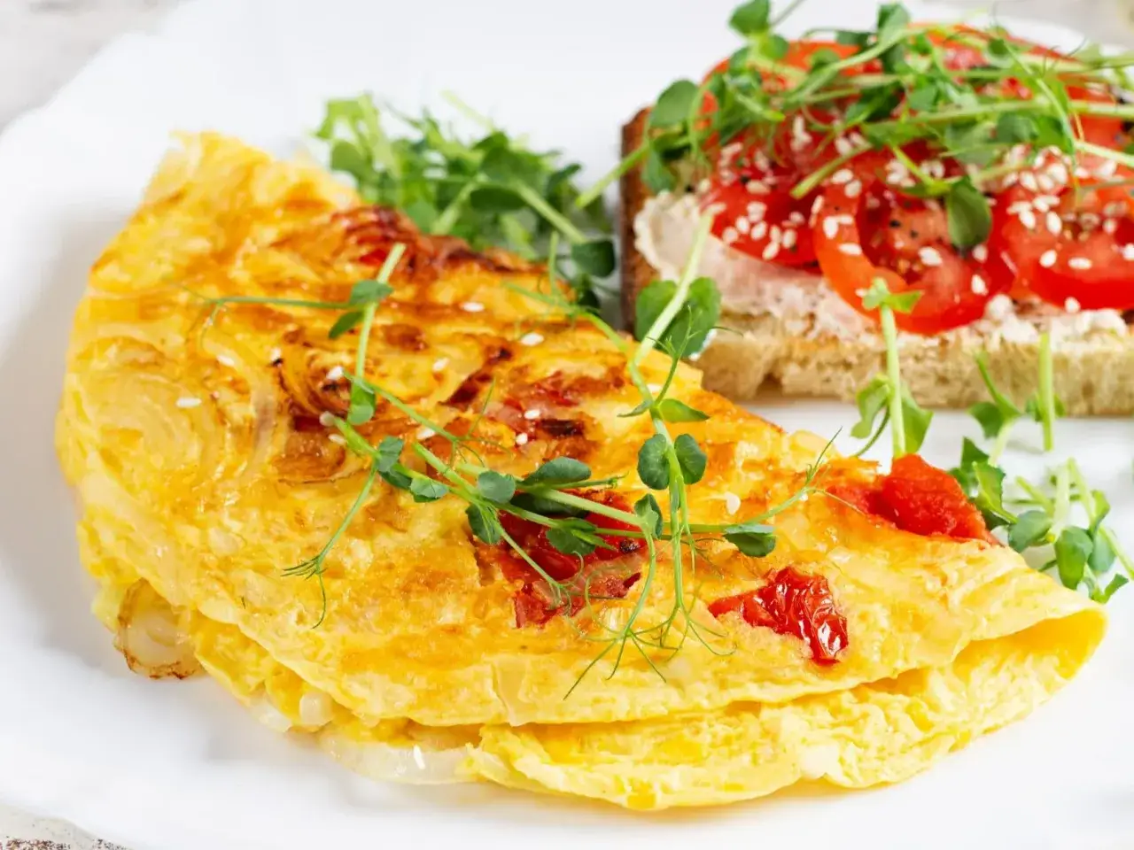 Złocisty omlet z pomidorami, udekorowany kiełkami, obok grzanki z pomidorami i kiełkami.