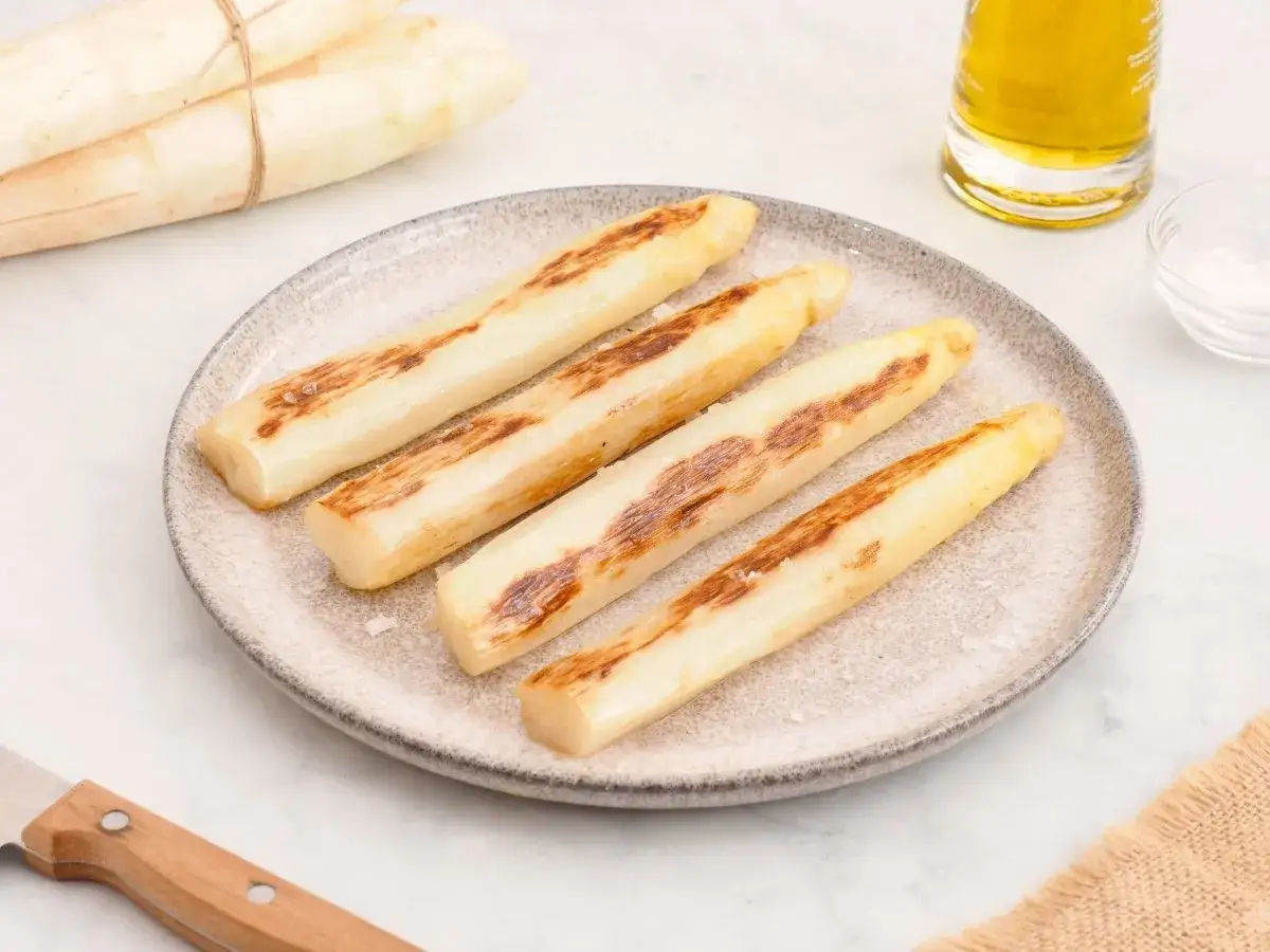 Cuatro esp&aacute;rragos blancos a la parrilla, con marcas doradas, reposan en un plato. Un manojo de esp&aacute;rragos blancos frescos yace al fondo.