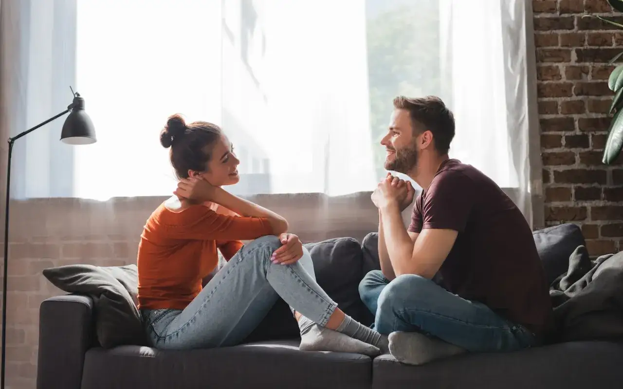 Coppia seduta sul divano, che parla e sorride. Un momento di complicità che fa pensare a come scopare bene, con intesa e passione.