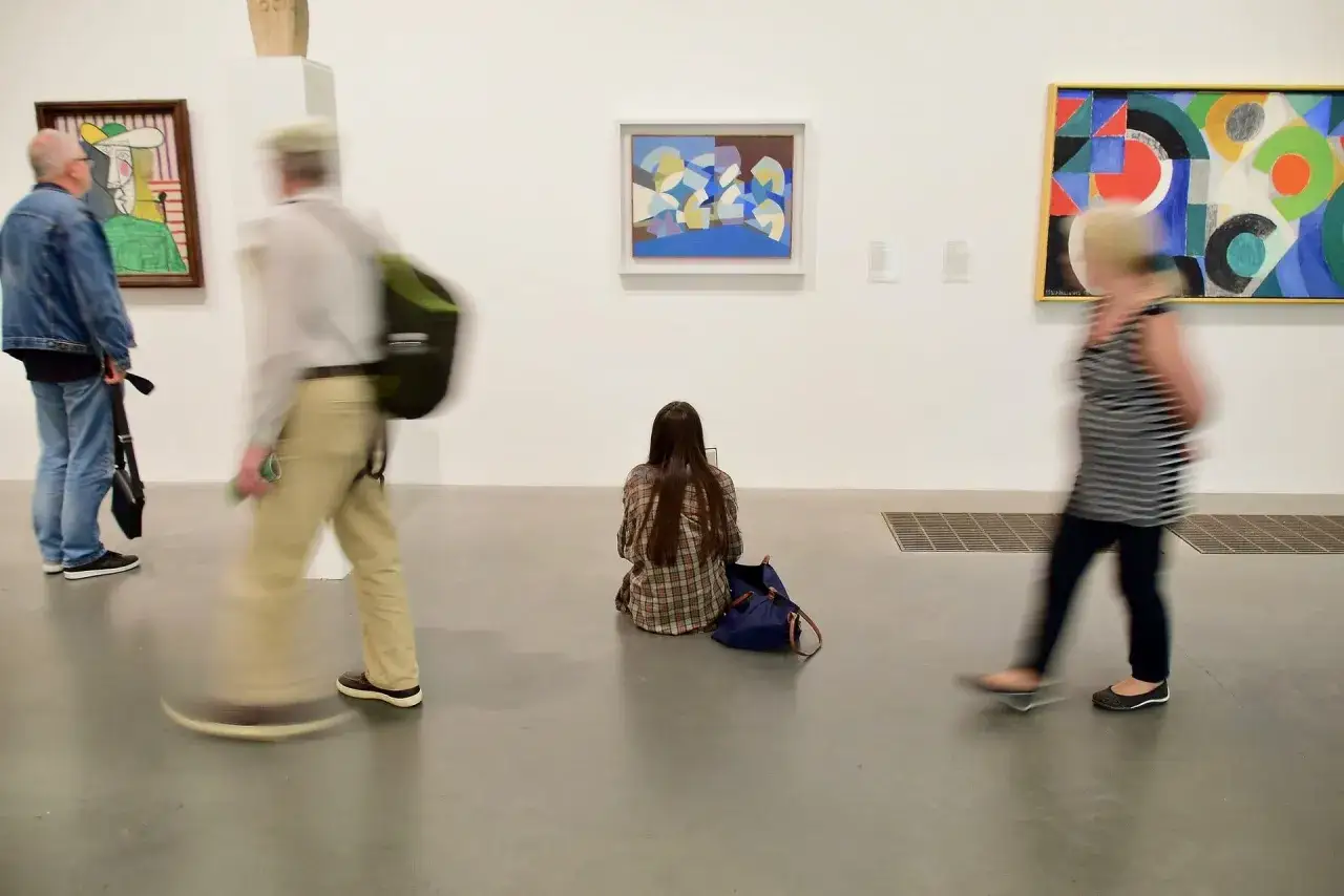 Visiteurs observant des œuvres d'art contemporain, une jeune femme assise au sol pour une analyse approfondie.