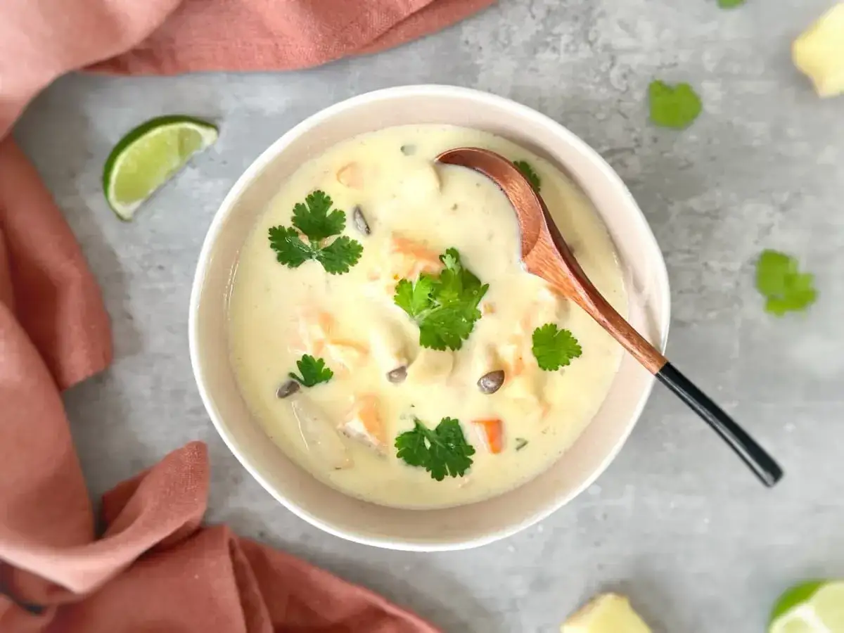 Un bol de blanquette de poisson crémeuse, garnie de coriandre fraîche et d'une cuillère en bois. Des quartiers de citron vert et des morceaux de gingembre sont disposés autour.