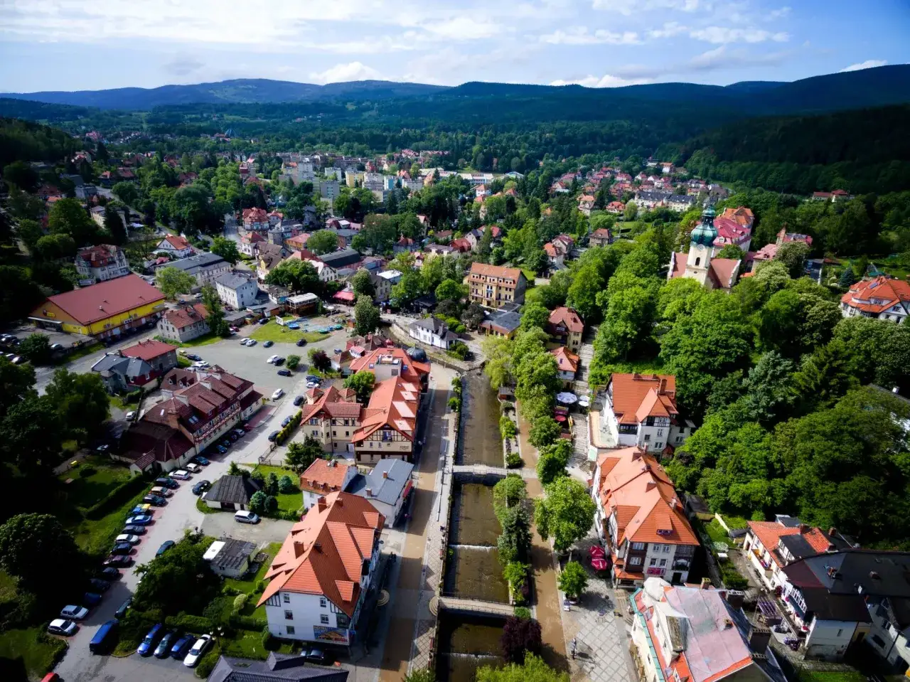 Widok z lotu ptaka na Polanicę-Zdrój, miasteczko z czerwonymi dachami i zielonymi wzgórzami w tle.
