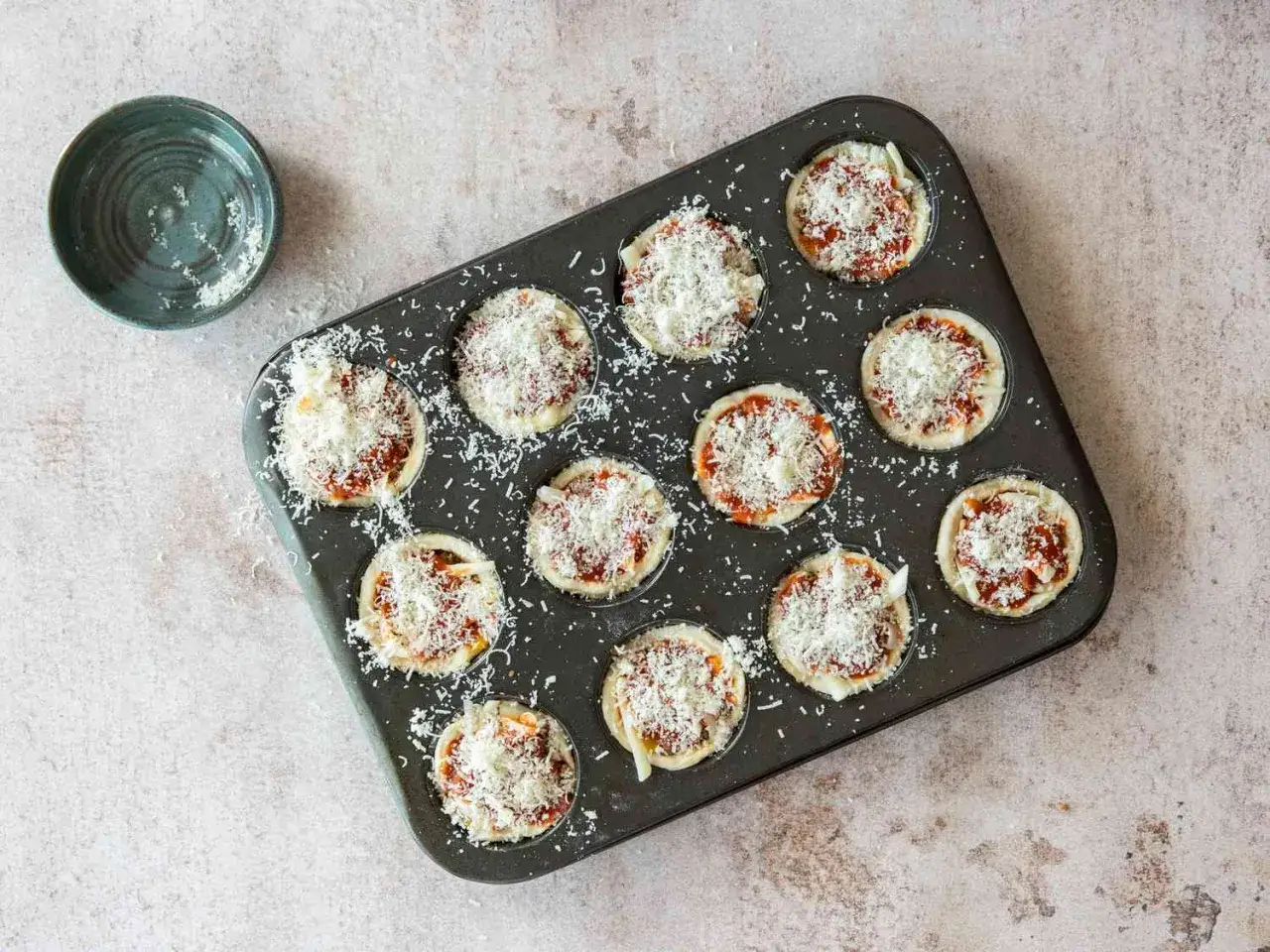 Zwölf kleine Pizza Muffins in einer Muffinform, bereit zum Backen, mit geriebenem Käse bestreut.