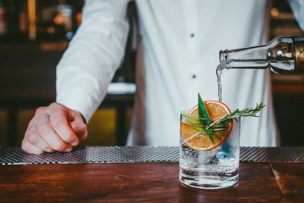 En bartender häller upp en gin sour i ett glas med is, apelsinskiva och rosmarin.