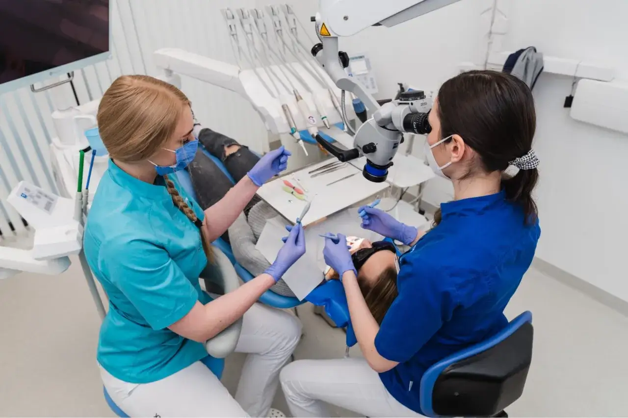 Kurs asystentka stomatologiczna: dwie kobiety w maseczkach i rękawiczkach pracują przy pacjencie, jedna używa mikroskopu.