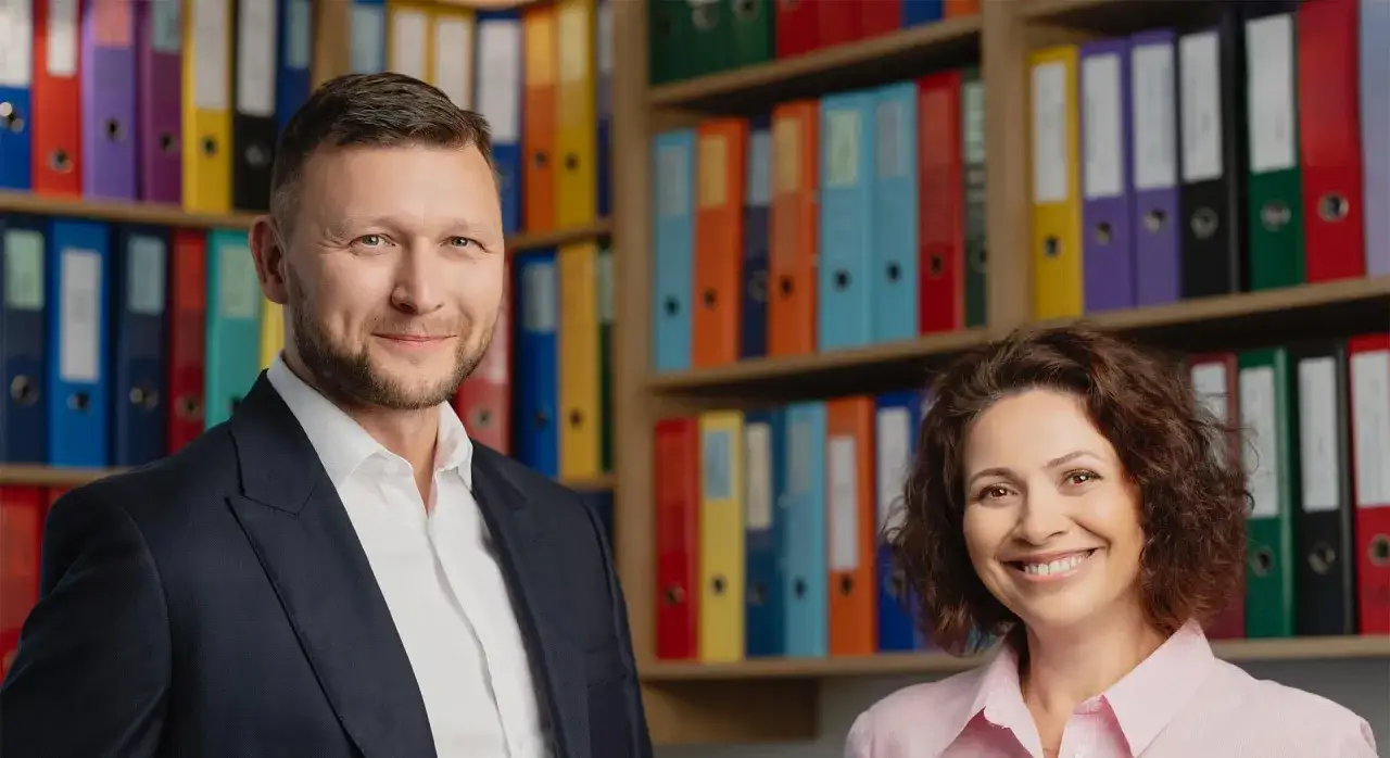 Uśmiechnięci mężczyzna i kobieta na tle regałów z kolorowymi segregatorami.