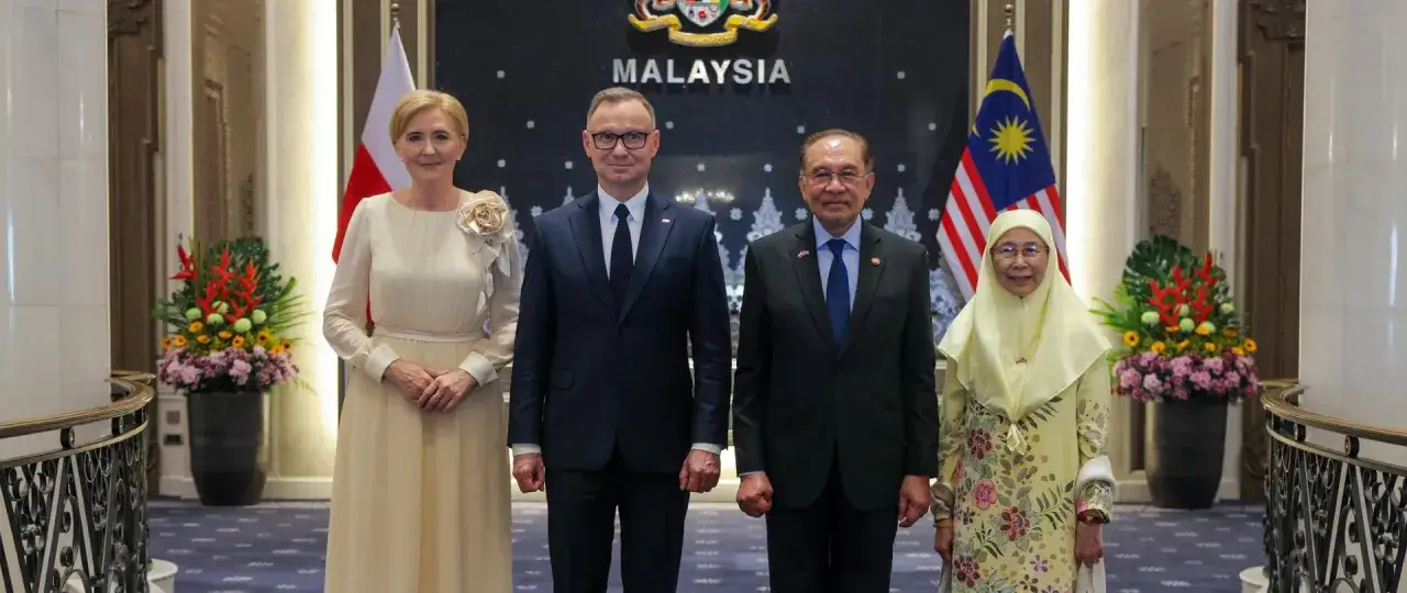 Pierwsza dama Polski w eleganckiej sukni obok prezydenta, na tle flag Malezji i godła państwowego.