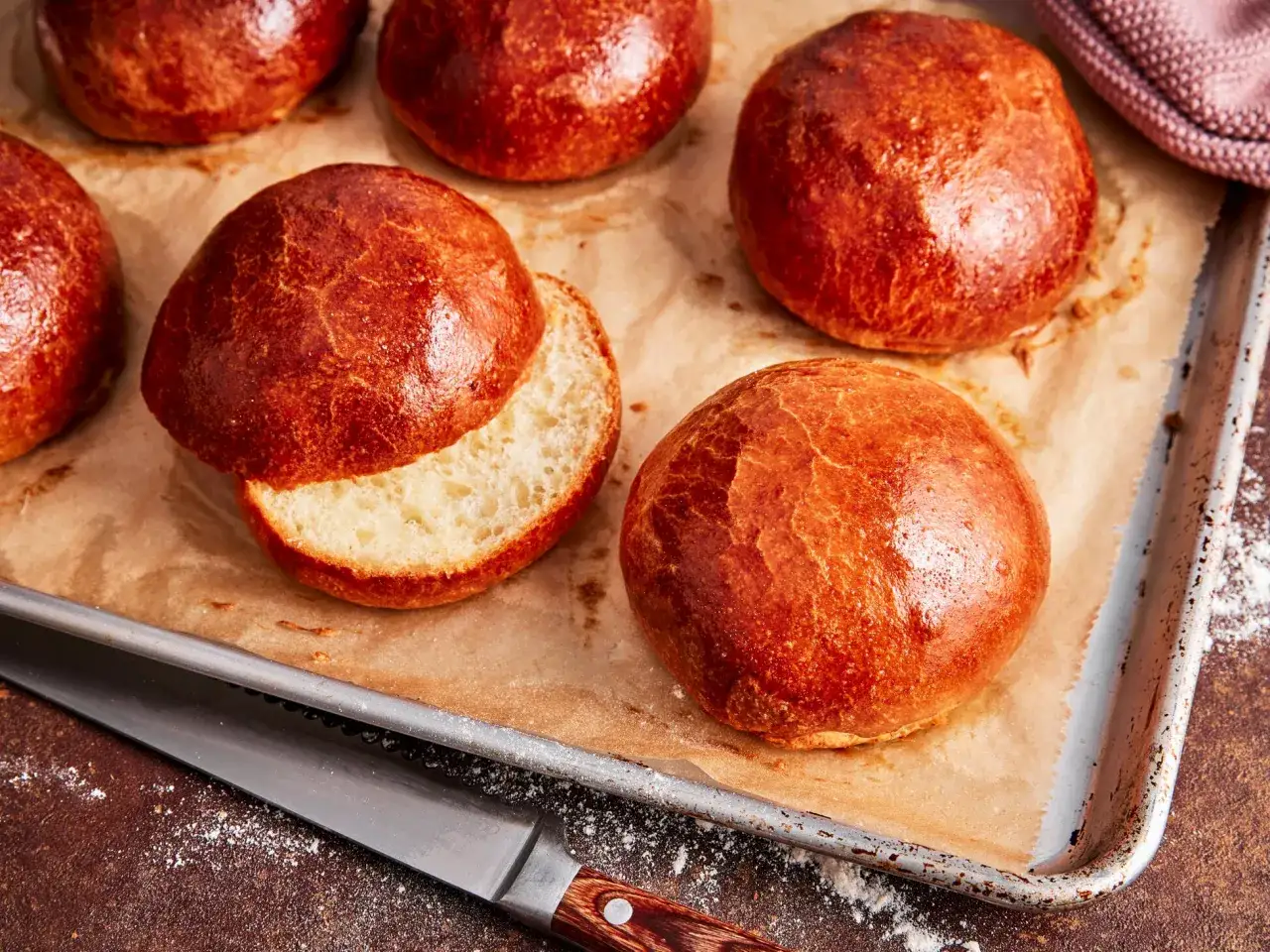 Frisch gebackene, goldbraune Brioche-Brötchen auf einem Backblech. Eines ist aufgeschnitten und zeigt die weiche Krume.