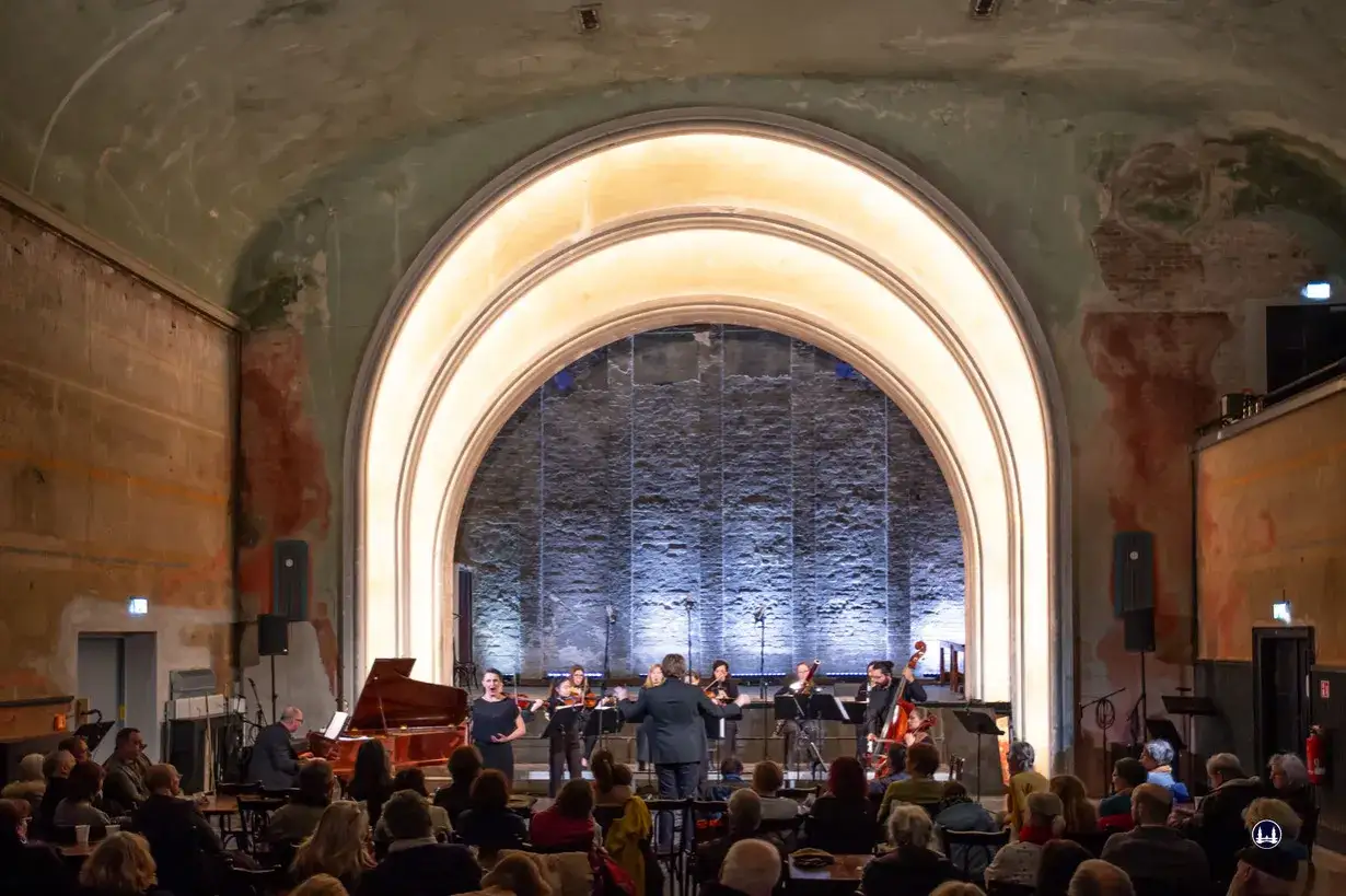 Ein Orchester spielt im **Theater im Delphi** vor einem Publikum. Eine S&auml;ngerin steht am Fl&uuml;gel, w&auml;hrend Musiker mit Streichinstrumenten und ein Cellist auf der B&uuml;hne agieren.