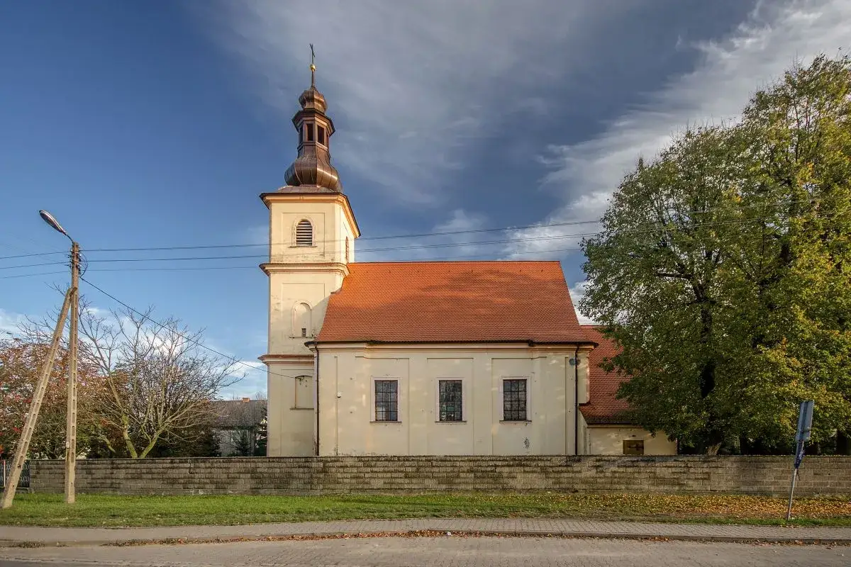 Jasny kościół z wieżą i czerwoną dachówką w Miłoszycach, otoczony murem i drzewami.