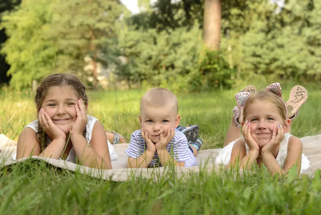 Trójka uśmiechniętych dzieci leży na kocu w trawie. Idealny przykład, jak ubrac dziecko na sesje zdjeciowa: jasne, letnie stroje.