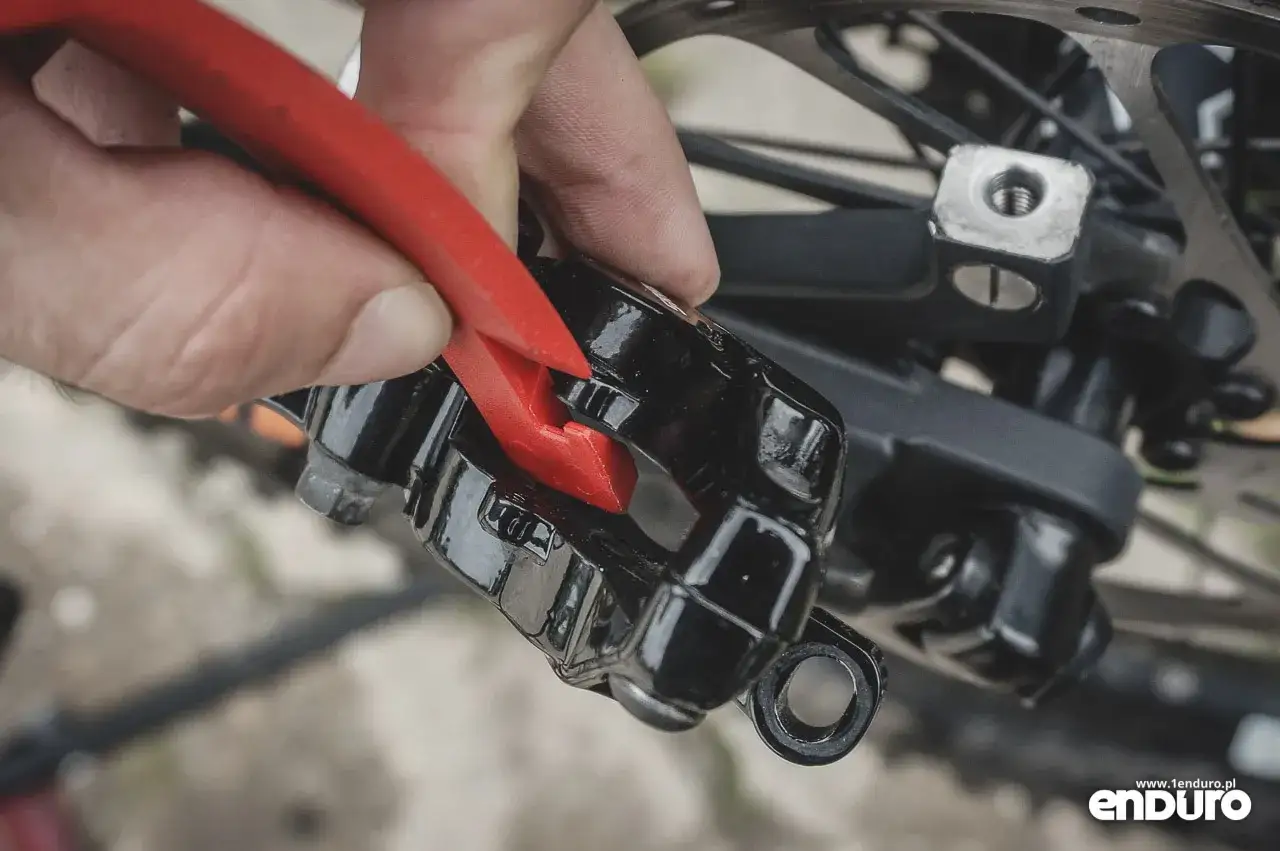 Dłoń z czerwonym narzędziem rozpycha tłoczek hamulca tarczowego roweru, przygotowując go do wymiany klocków. Poradnik: jak wymienić klocki hamulcowe w rowerze.