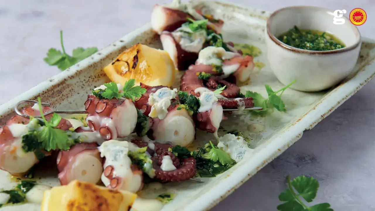 Tentacoli di polpo alla griglia su spiedini, conditi con erbe fresche, formaggio e limone, serviti con salsa verde.
