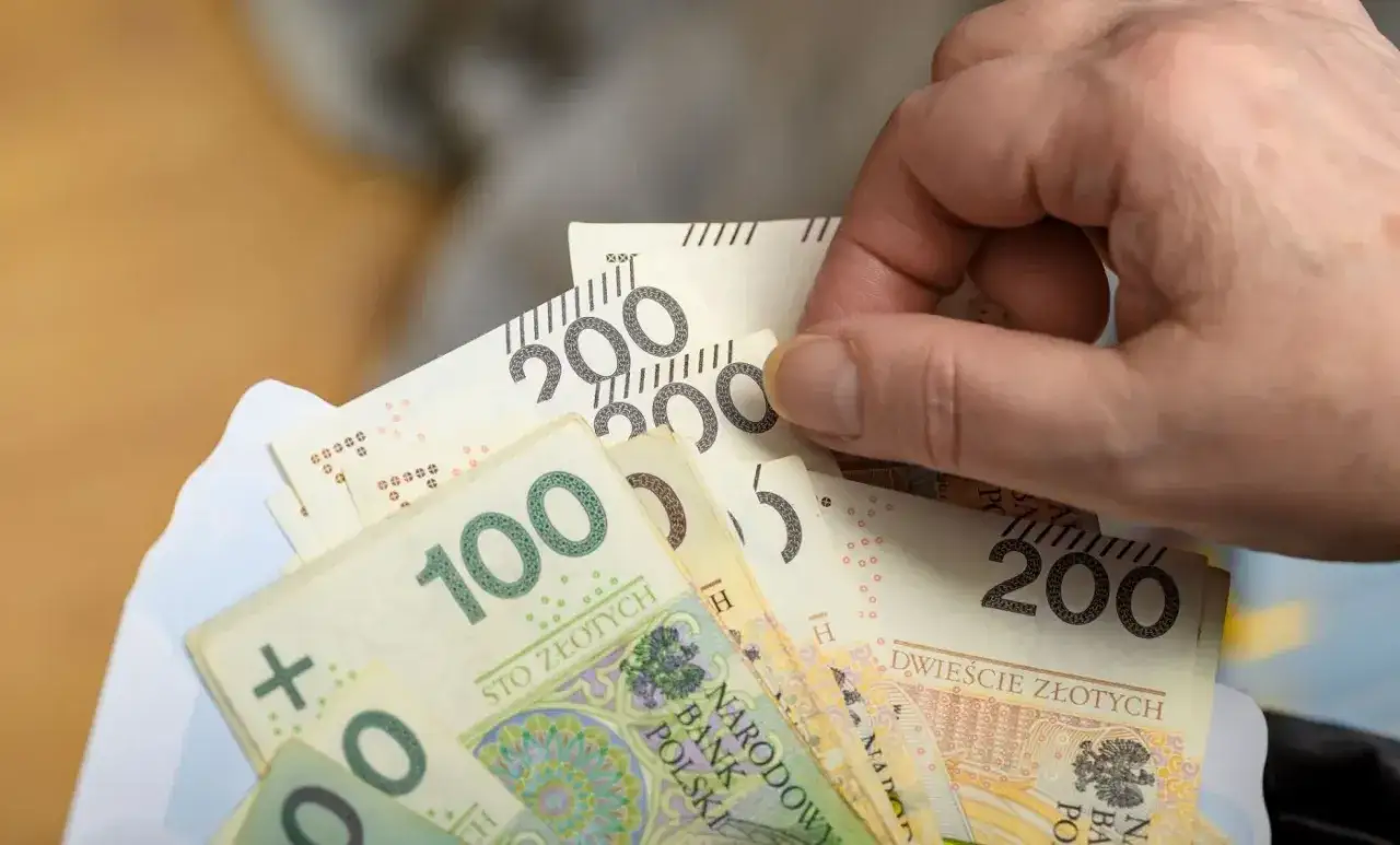 Dłoń trzyma plik banknotów 100 i 200 zł. Może to symbolizować drugą waloryzację emerytur i wzrost dochodów.