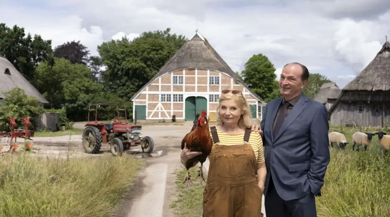 Ein Mann im Anzug und eine Frau mit Huhn vor einem Bauernhaus. Ein Traktor und Schafe im **weiten Land** auf einem Hof, wie aus der **Mediathek**.
