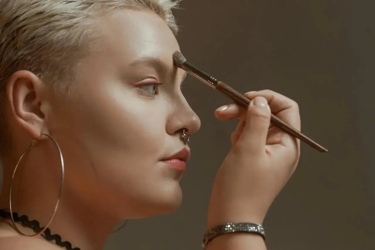 Kobieta z krótkimi blond włosami i kolczykiem w nosie nakłada bronzer na czoło pędzlem.