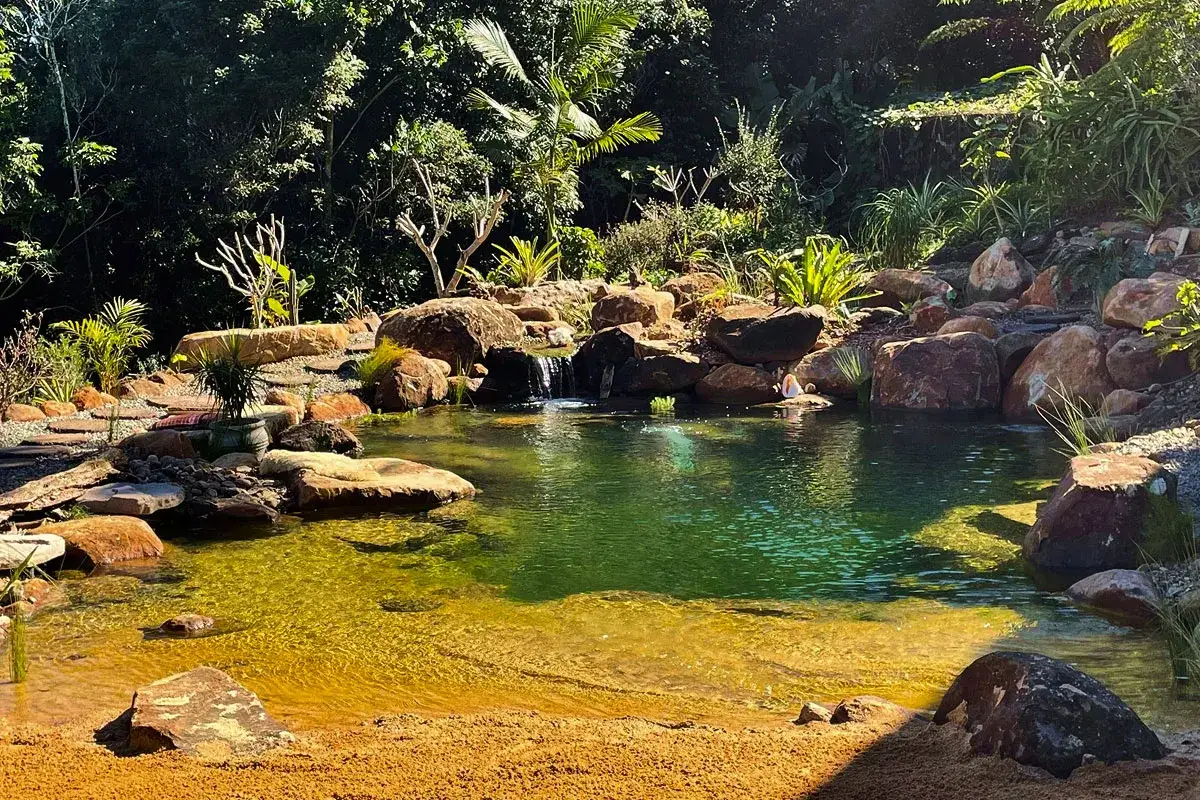 En vacker natur pool med ett litet vattenfall, omgiven av stenar och frodig grönska. En person sitter vid kanten.