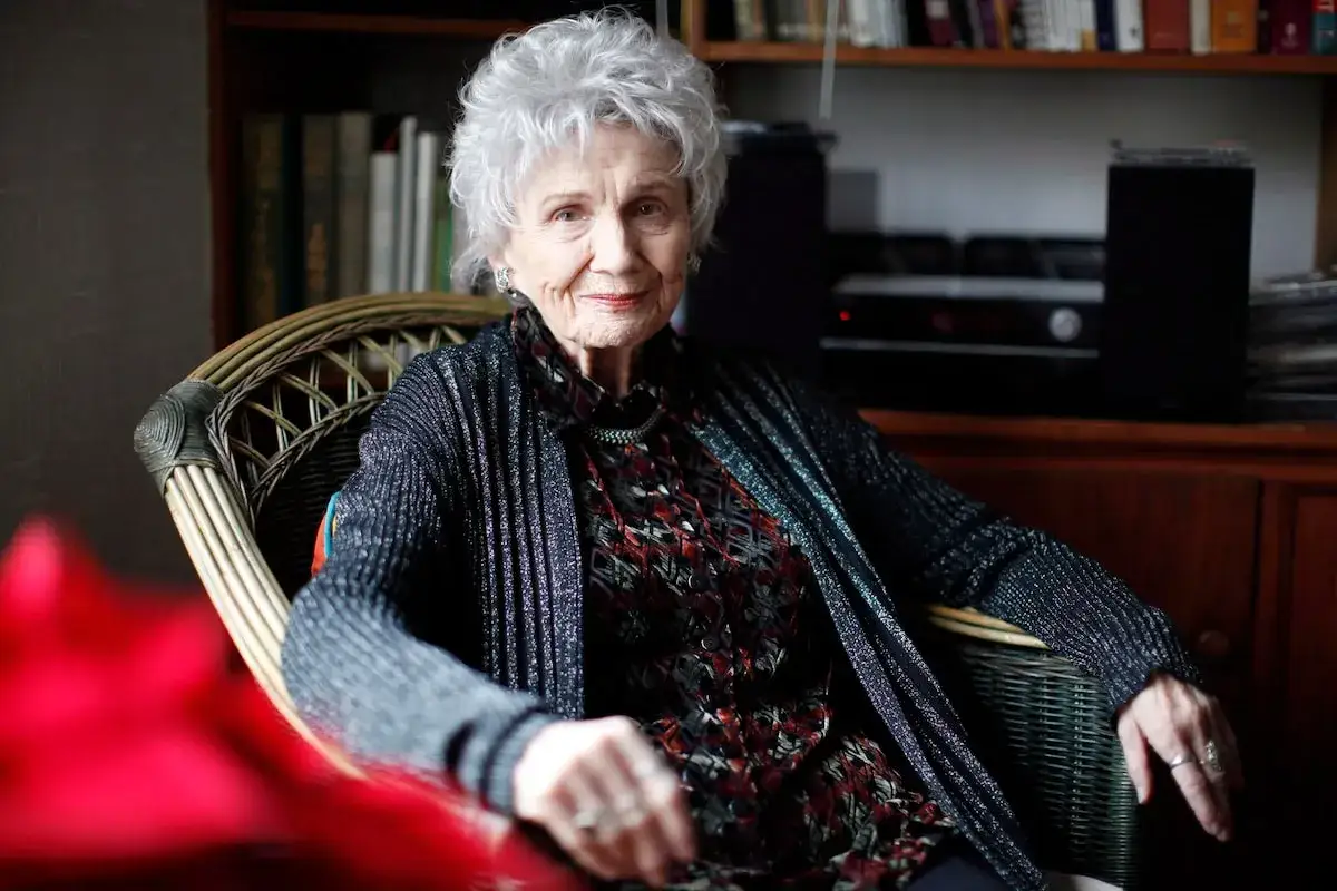 Ritratto di Alice Munro, con i suoi capelli bianchi e un sorriso gentile, seduta su una sedia di vimini.