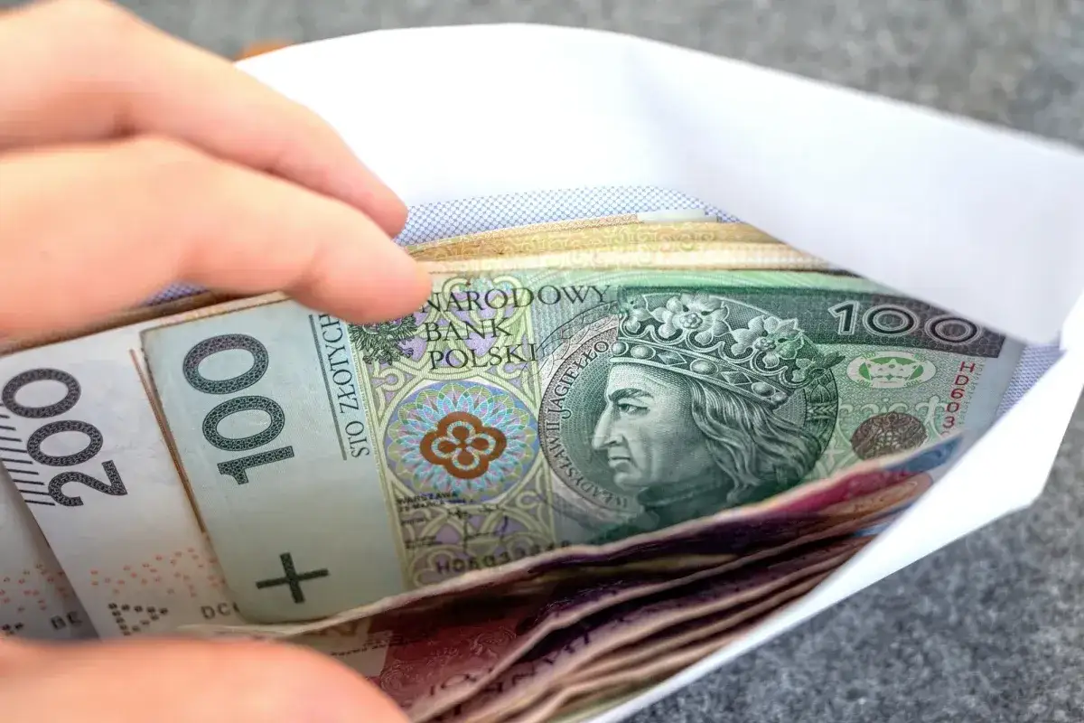 Dłoń wsuwa kopertę z banknotami 100 i 200 zł. Pieniądze mogą symbolizować wsparcie, np. renta wdowia komu przysługuje.