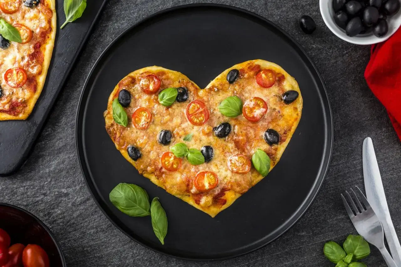 Pizza walentynkowa w kształcie serca, ozdobiona pomidorkami, oliwkami i bazylią, na czarnym talerzu.