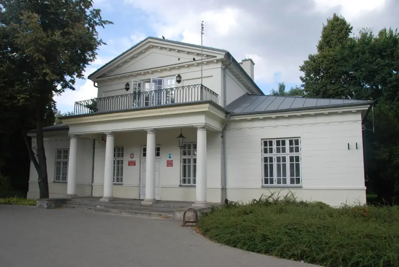 Pałac Potulickich w Pruszkowie, biały budynek z kolumnami i balkonem, otoczony zielenią.