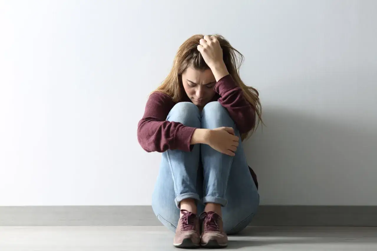Ragazza seduta a terra, abbraccia le ginocchia, testa china, capelli lunghi, maglione bordeaux, jeans chiari, scarpe sportive. Sembra triste, perche non so che dire.