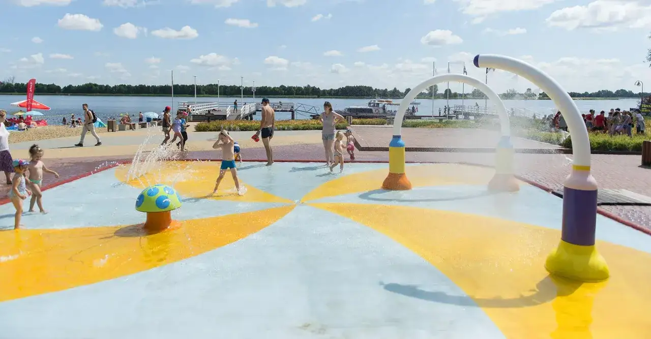 Kolorowe **jezioro z atrakcjami dla dzieci** oferuje wodne szaleństwo. Dzieci pluskają się w fontannach, a w tle widać plażę i spokojną taflę wody.