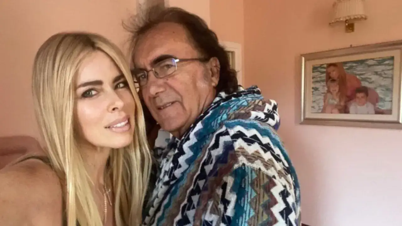 Al Bano i jego nowa partnerka, uśmiechnięci, pozują do zdjęcia. W tle wisi obraz z rodziną.
