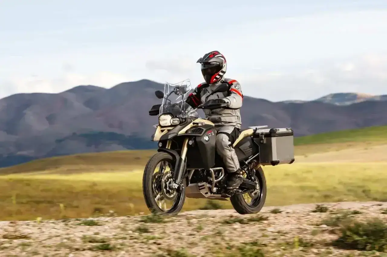 Motocyklista w kasku i stroju podr&oacute;żuje motocyklem BMW 800R po piaszczystej drodze, w tle g&oacute;ry.