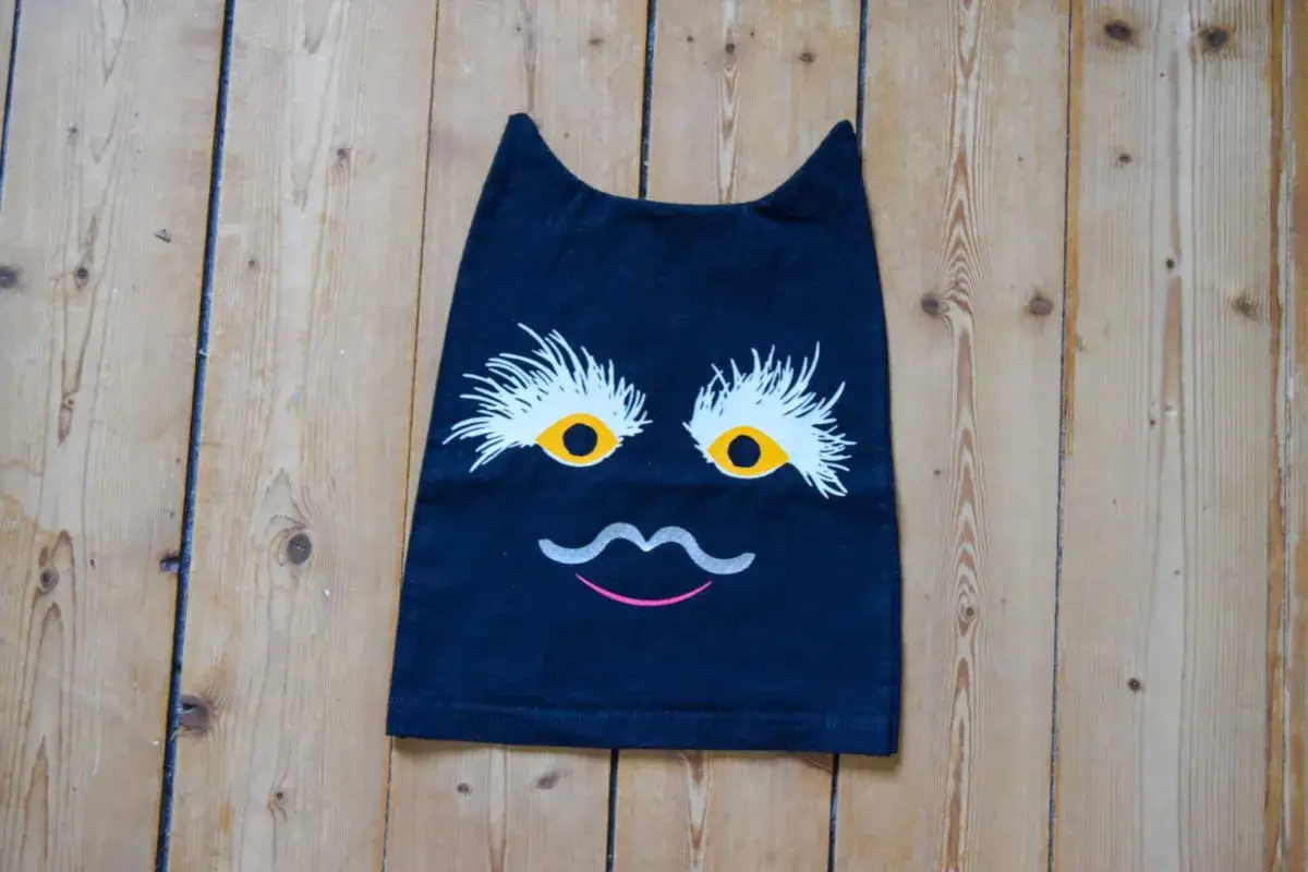 En svart tr&ouml;ja med ett roligt kattansikte, som om det vore en sy sommarskuggan. &Ouml;gonen &auml;r stora och gula med vita fransar, och en gr&aring; mustasch och rosa mun full&auml;ndar uttrycket.