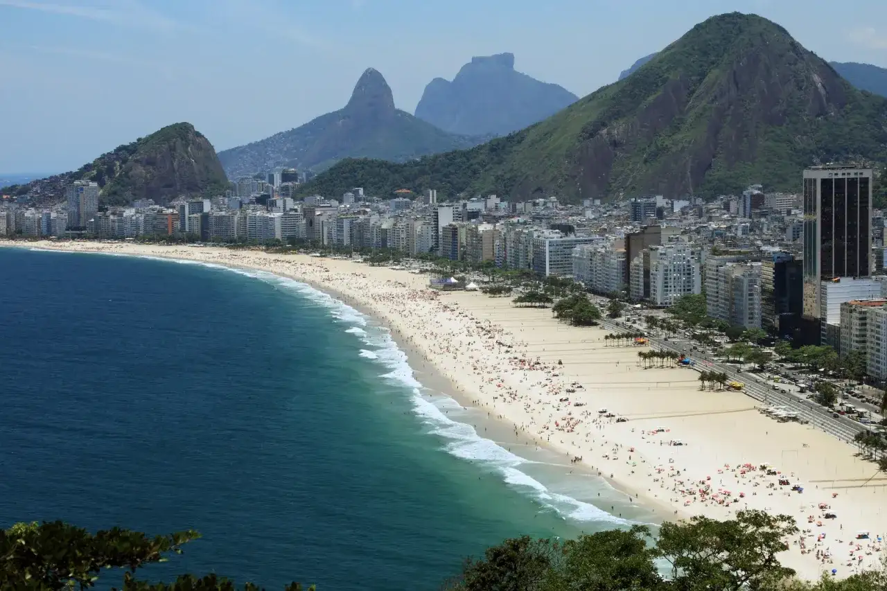 Słynna plaża w Rio de Janeiro tętni życiem. Tłumy ludzi odpoczywają na piasku, a fale obmywają brzeg. W tle widać góry i miasto.