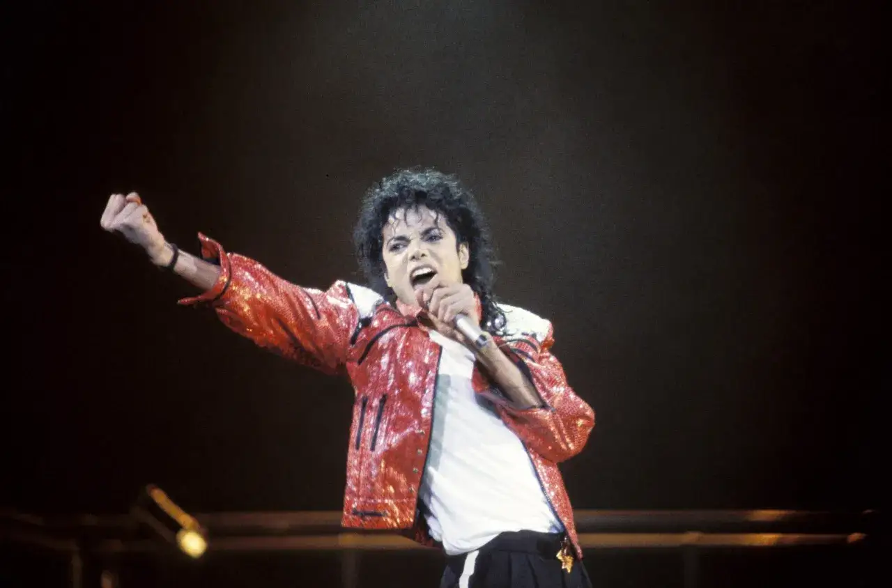 Kr&oacute;l popu, Michael Jackson, w błyszczącej czerwonej kurtce śpiewa na scenie, unosząc pięść w geście triumfu.