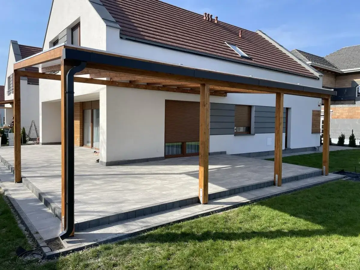 Nowy, tani ogród zimowy z drewnianą konstrukcją i nowoczesnym dachem, przylegający do białego domu z brązowymi roletami.