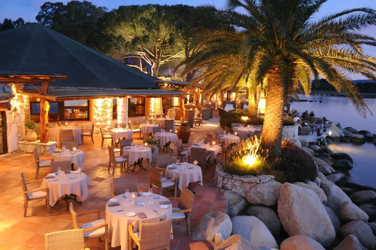 Terrasse illumin&eacute;e d'un restaurant chic au bord de l'eau, palmiers et tables dress&eacute;es. Le meilleur restaurant Porto Vecchio pour une soir&eacute;e magique.