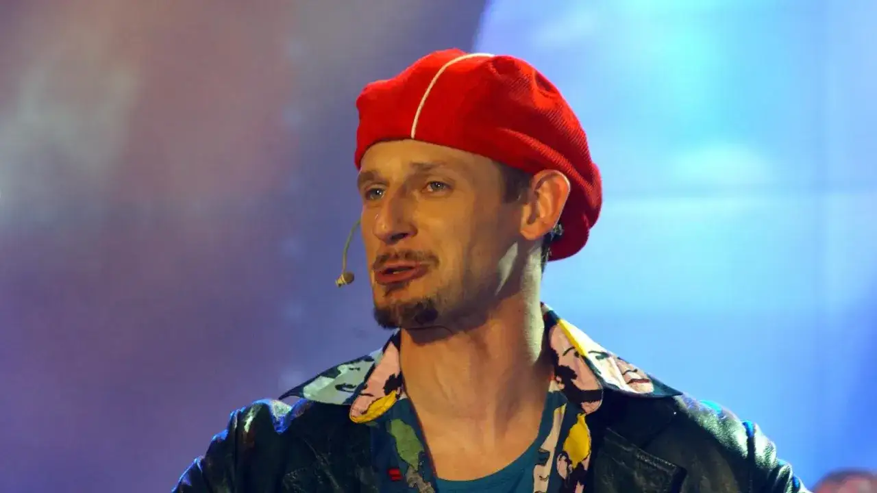 Michał Wójcik w czerwonej beretce i kolorowej koszuli, z mikrofonem przy uchu, na scenie.