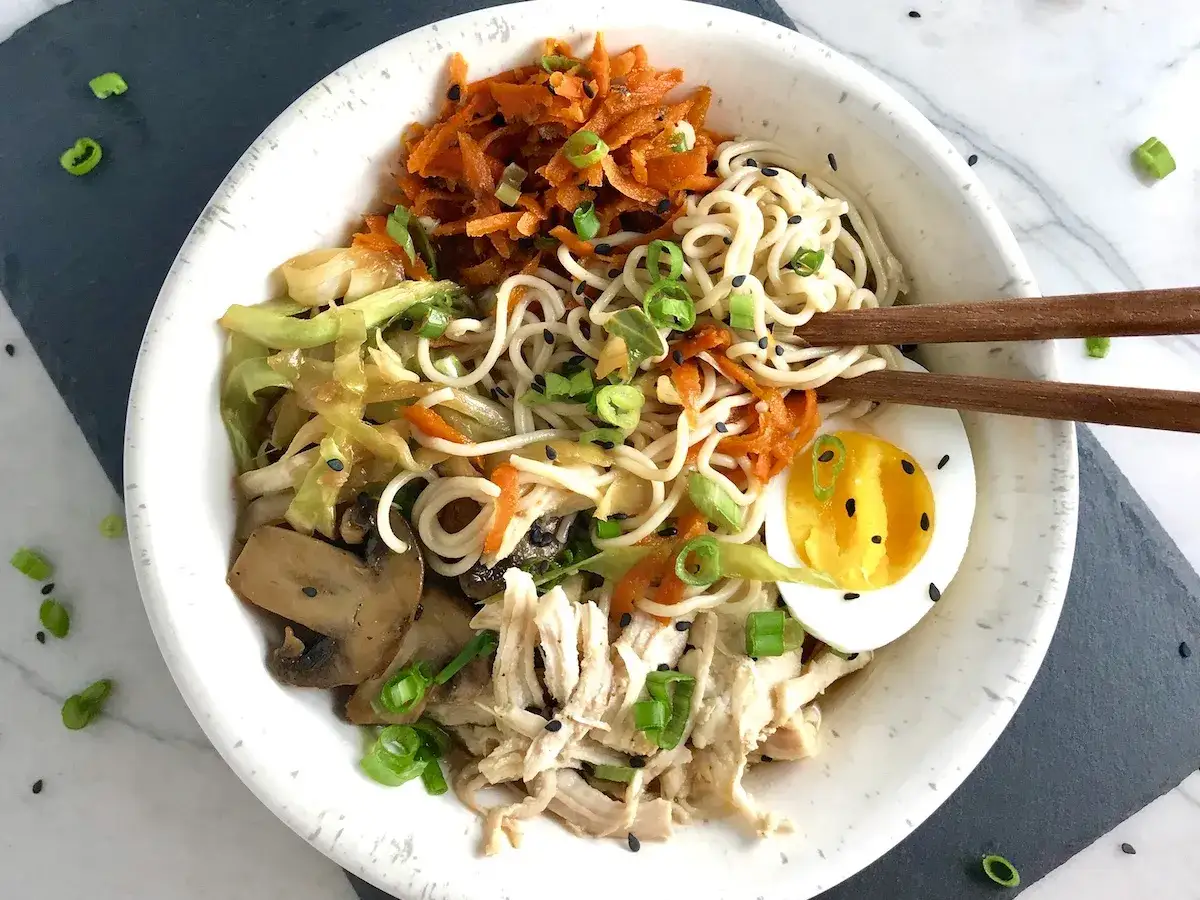 Un bol de ramen poulet savoureux avec nouilles, carottes râpées, champignons, chou, œuf mollet et oignons verts. Une véritable recette ramen poulet.