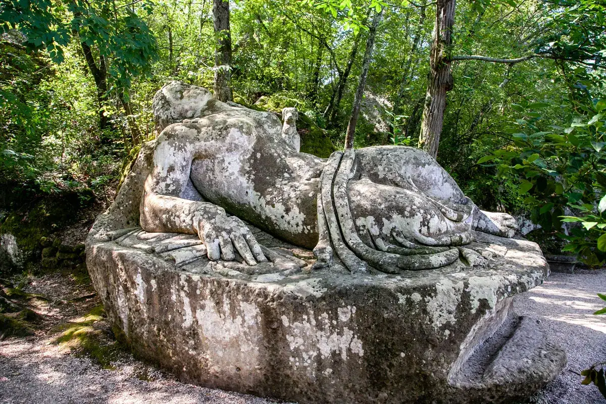 Gigantesca statua in pietra, ricoperta di muschio, giace nel sacro bosco. Un'antica figura mitologica, forse un titano, riposa tra la vegetazione lussureggiante.