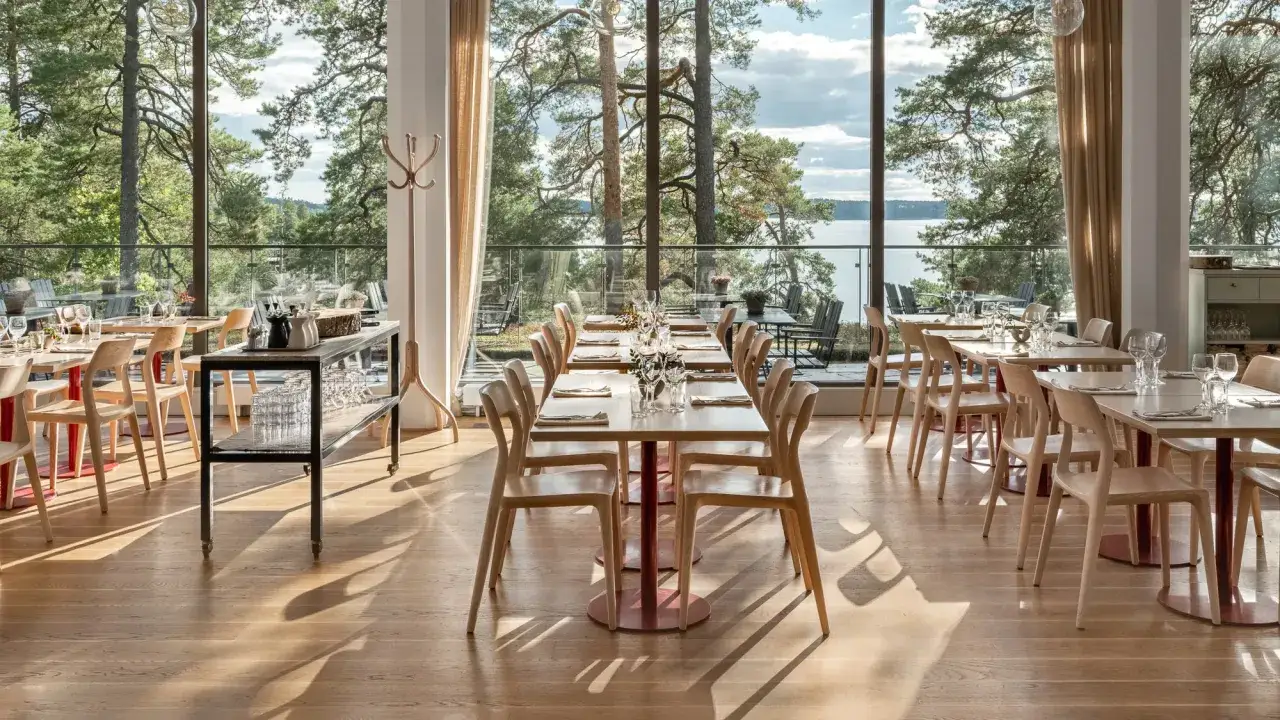 Vacker matsal med utsikt över vattnet och skogen på Artipelag Stockholm. Ljusa träbord och stolar, solljus som strömmar in.