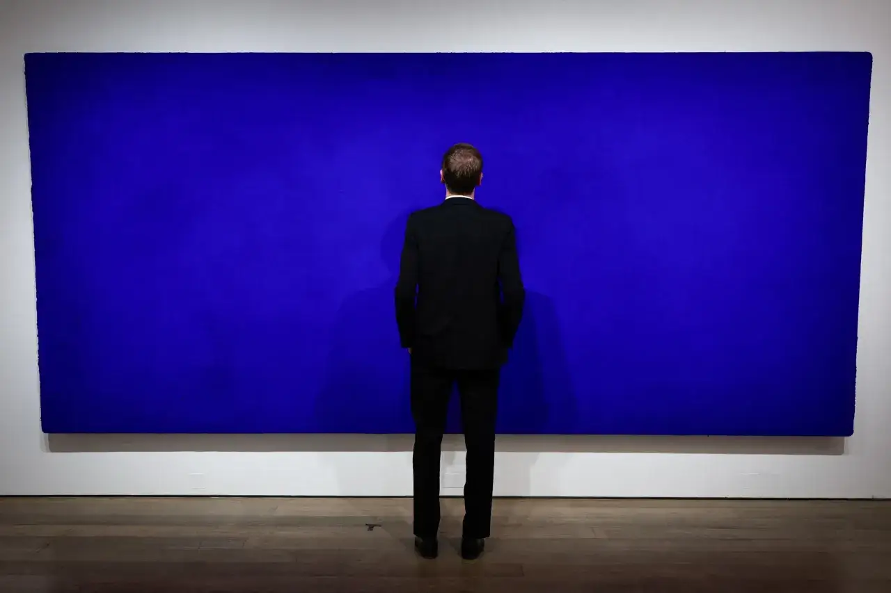 Un homme en costume noir contemple une immense peinture monochrome bleue, absorbé par sa profondeur.