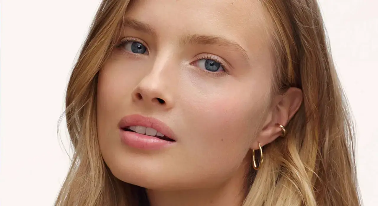 Visage d'une jeune femme aux yeux bleus, peau lumineuse, lèvres rosées. Un style maquillage no makeup, rehaussé par des boucles d'oreilles dorées.