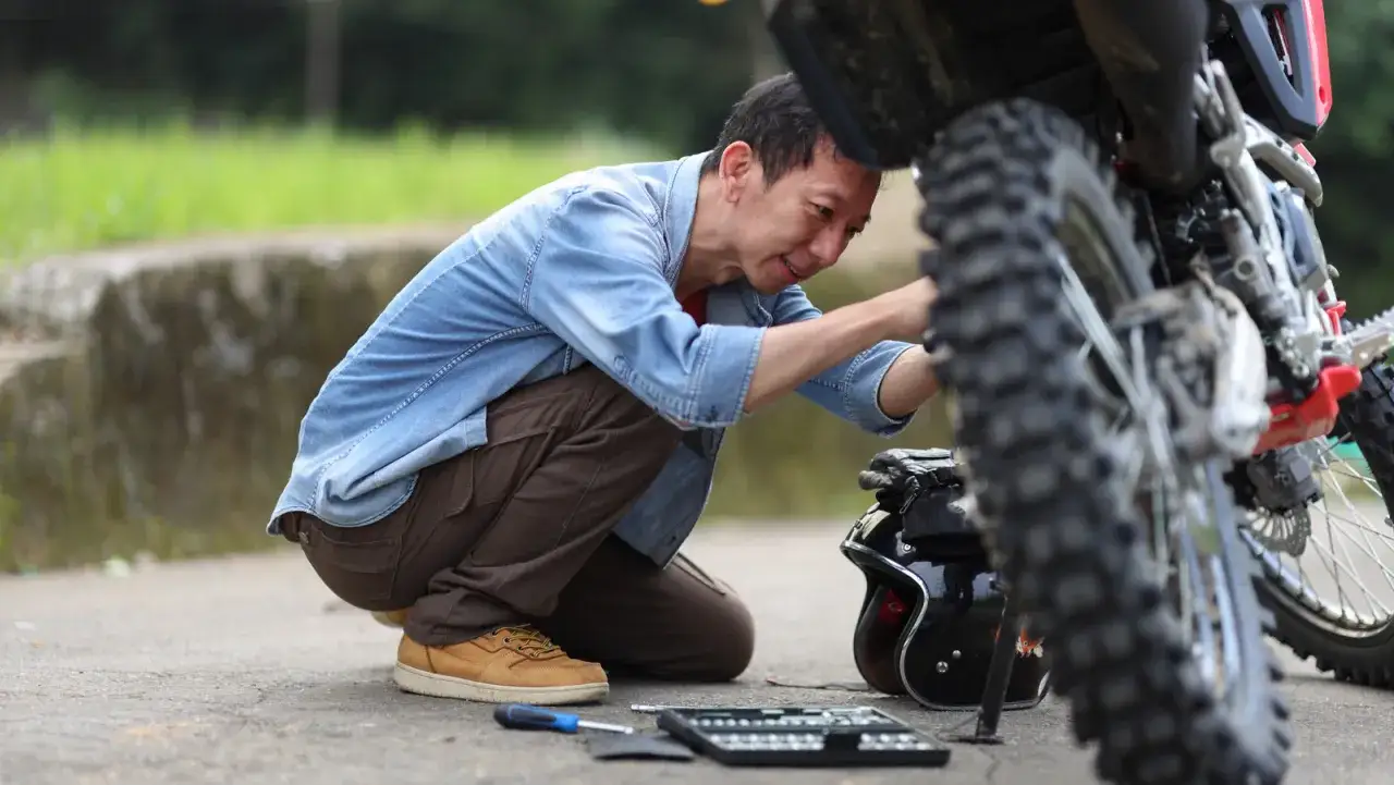 Uomo con attrezzi cerca di capire perch&eacute; il motore non parte, inginocchiato accanto alla sua moto.