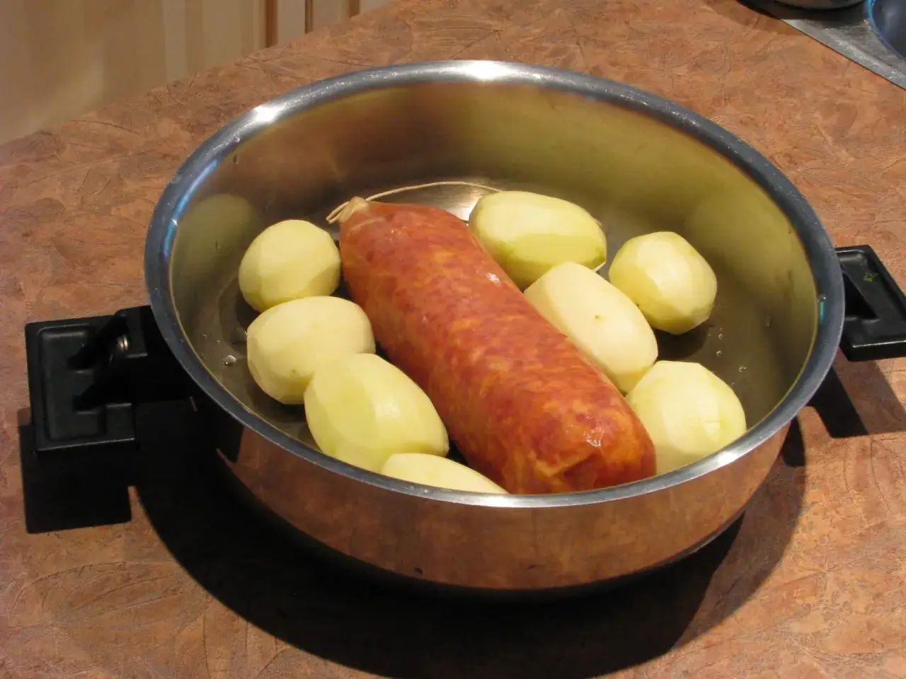 Un saucisson est prêt à être cuit avec des pommes de terre pelées dans une casserole en métal.