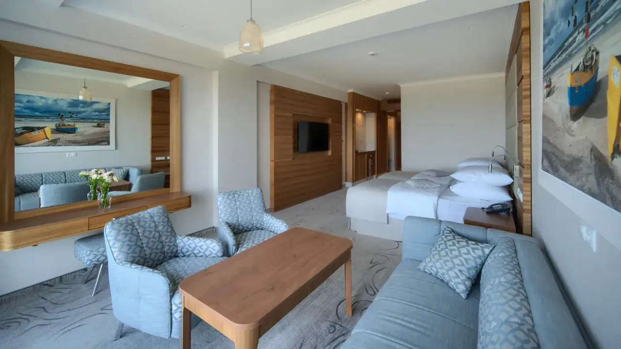 Pokój w hotelu Gołębiewski Morze z dwoma łóżkami, sofą, fotelami i stolikiem. W tle obraz z łodziami na plaży.