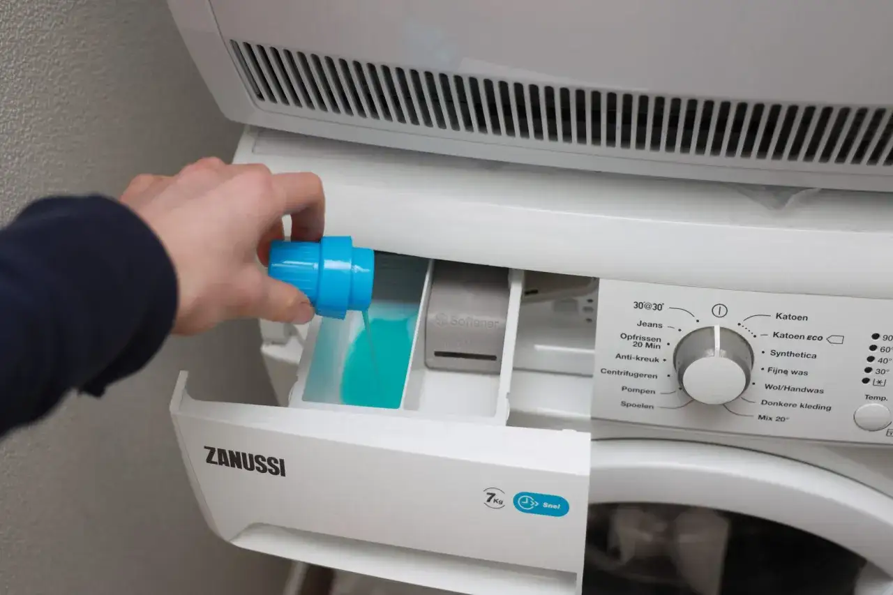 Hand giet blauw wasmiddel in het bakje van de wasmachine. Links of rechts, het bakje voor wasmiddel is duidelijk zichtbaar.