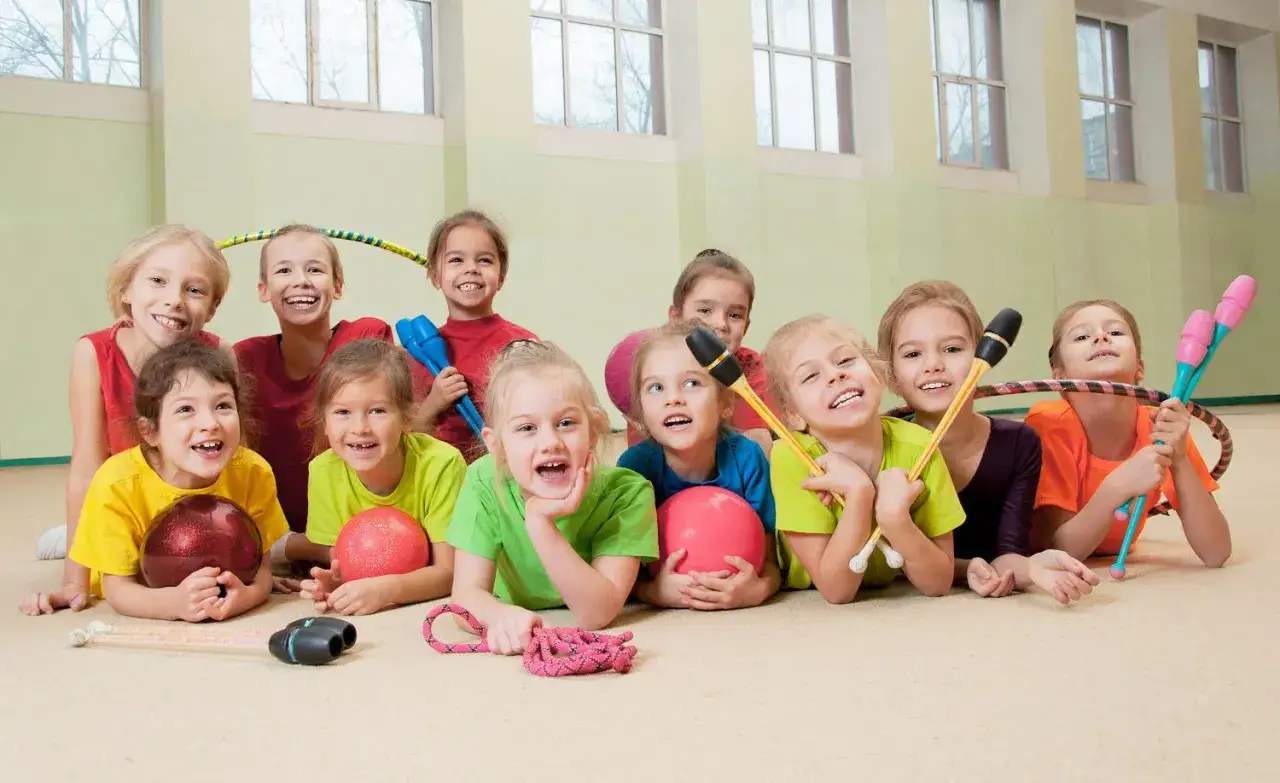 Fröhliche Kinder liegen auf dem Boden einer Turnhalle, bereit für Bewegungsspiele in der Grundschule. Sie halten Bälle, Hula-Hoops und Keulen.