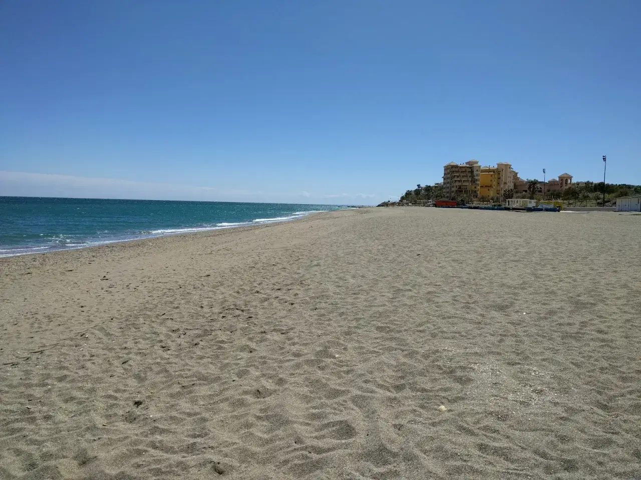 Ein weiter, sonniger fuengirola strand mit ruhigem Meer und einigen Geb&auml;uden im Hintergrund.