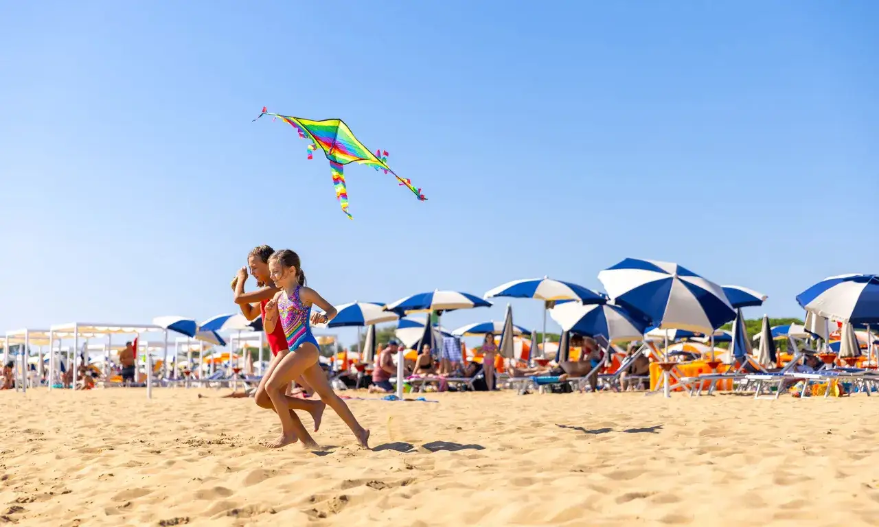 Dwie dziewczynki biegną po piaszczystej plaży w Bibione, puszczając kolorowego latawca. W tle widać parasole i leżaki.