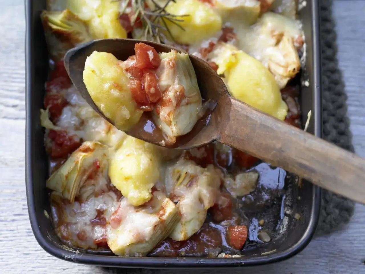 Gnocchi alla Romana rezept: herzhafte Gnocchi mit Artischocken, Tomaten und geschmolzenem K&auml;se in einer Auflaufform, serviert mit einem Holzl&ouml;ffel.